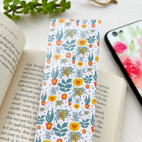 Hand holding a floral patterned bookmark over an open book with a phone and plant in the background.