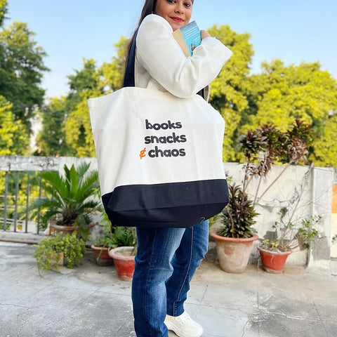 Woman carrying large canvas tote bag that says books, snacks & chaos.