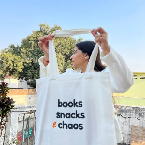 Woman holding white canvas tote bag with “Books Snacks & Chaos” printed on it outdoors