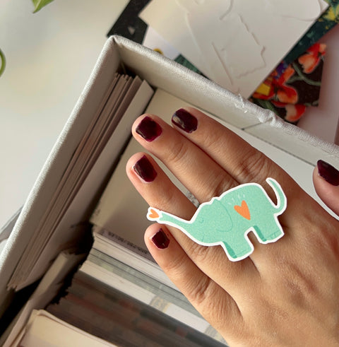 Hand wearing a green elephant-shaped ring with a heart on a blurred background