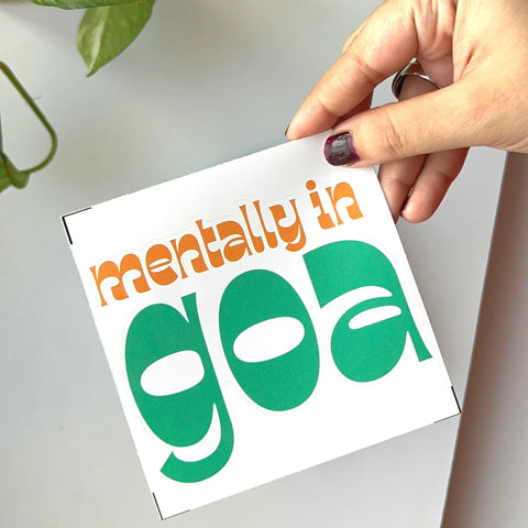 Hand holding a 'Mentally in Goa' laptop sticker with retro typography and travel vibes