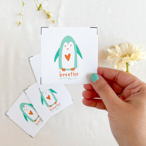 Hand holding a card with a penguin illustration and 'breathe' text on a light surface with flowers.