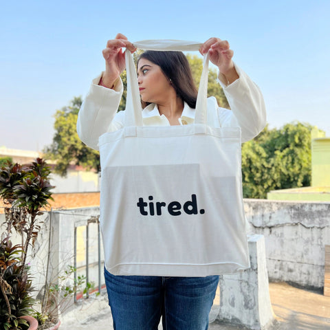 White canvas tote bag with bold “tired.” text print, held by a person with dark red nail polish